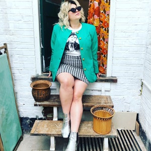 May include: A woman wearing a green blazer, a white t-shirt with a black and white graphic print, a black and white checked skirt, and silver ankle boots. She is sitting on wooden steps with a wicker basket on each step.