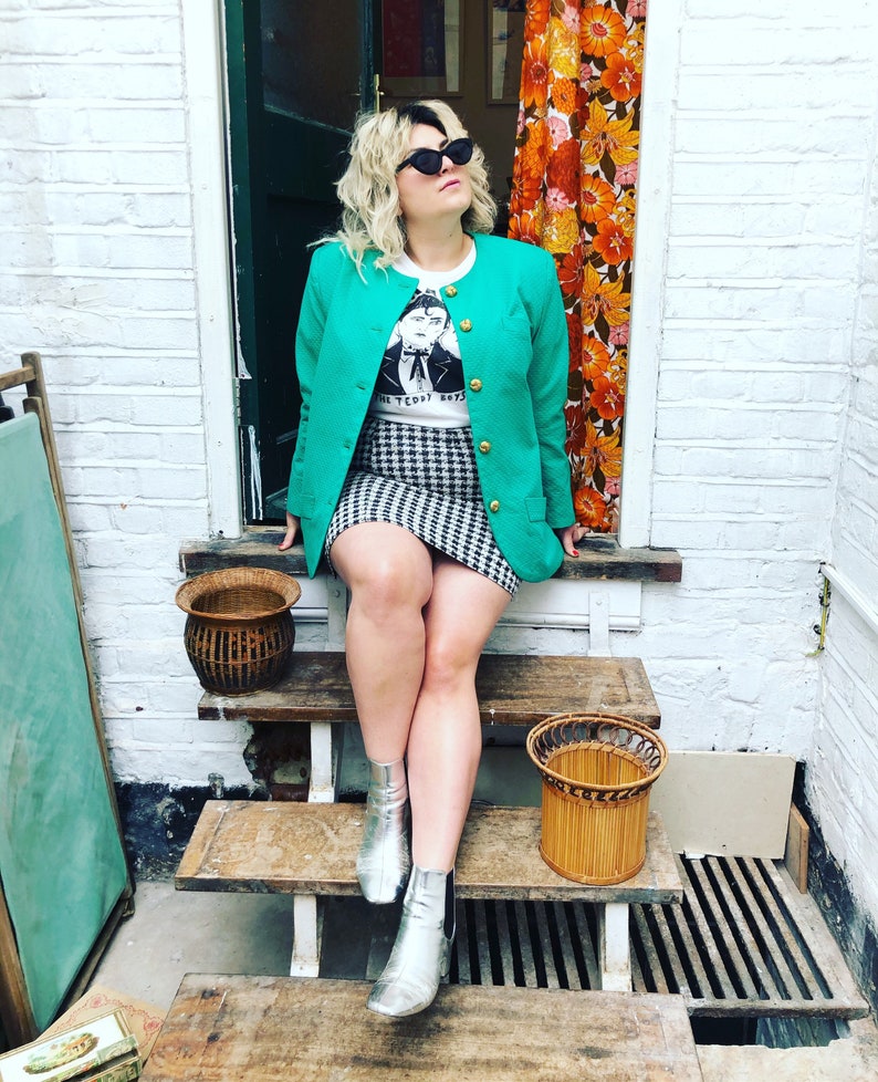 May include: A woman wearing a green blazer, a white t-shirt with a black and white graphic print, a black and white checked skirt, and silver ankle boots. She is sitting on wooden steps with a wicker basket on each step.