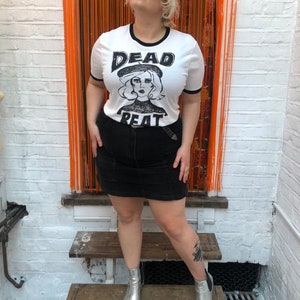 May include: A white t-shirt with a black ringer and a black and white graphic print of a woman wearing a beret with the text "DEAD BEAT" on the front. The person wearing the shirt is standing on a wooden staircase and wearing a black skirt and silver boots.