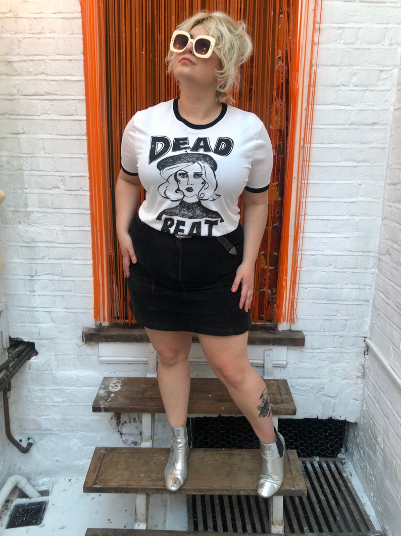 May include: A white t-shirt with a black ringer and a black and white graphic print of a woman wearing a beret with the text "DEAD BEAT" on the front. The person wearing the shirt is standing on a wooden staircase and wearing a black skirt and silver boots.