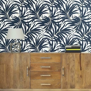 Pu&ograve; includere: Una credenza in legno con quattro cassetti e un'anta. Una lampada argentata con un paralume bianco &egrave; appoggiata sopra la credenza. Lo sfondo &egrave; una carta da parati bianca con un motivo a foglie di palma blu navy.