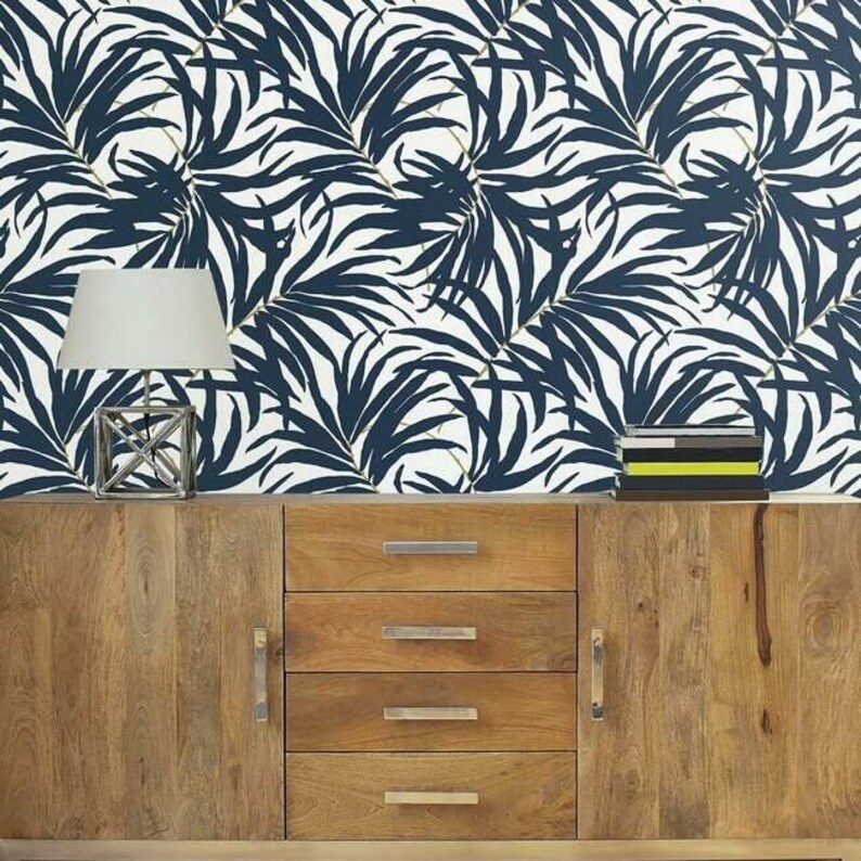 Pu&ograve; includere: Una credenza in legno con quattro cassetti e un'anta. Una lampada argentata con un paralume bianco &egrave; appoggiata sopra la credenza. Lo sfondo &egrave; una carta da parati bianca con un motivo a foglie di palma blu navy.
