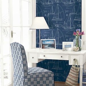 Op de afbeelding: Een wit bureau met een blauw-wit gepatroneerde stoel ervoor. Op het bureau staan een witte lamp, een laptop en een vaas met bloemen. De muur achter het bureau is bedekt met blauwe behang met een wit zeilbootpatroon.