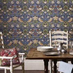 May include: A dining room with a wallpaper featuring a repeating pattern of birds, flowers, and leaves in shades of blue, green, and brown. Two white chairs with a red and green patterned pillow on one chair are placed on either side of a wooden table with a stack of white plates, a silver pitcher, and two glasses.