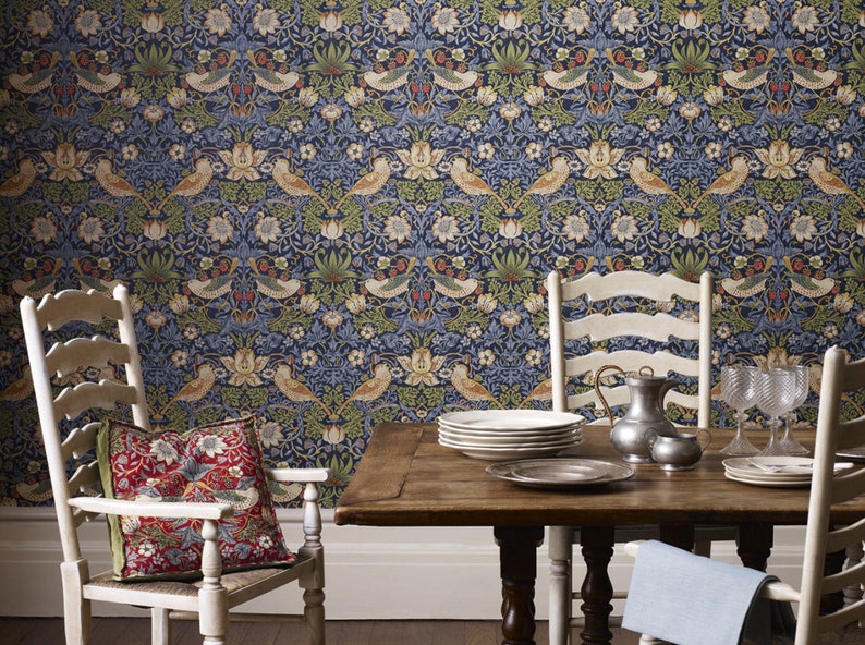 May include: A dining room with a wallpaper featuring a repeating pattern of birds, flowers, and leaves in shades of blue, green, and brown. Two white chairs with a red and green patterned pillow on one chair are placed on either side of a wooden table with a stack of white plates, a silver pitcher, and two glasses.