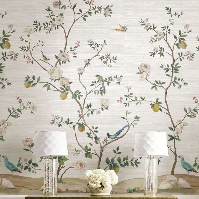 K&ouml;nnte beinhalten: Eine wei&szlig;e Tapete mit einem floralen Muster, das ein sich wiederholendes Design aus &Auml;sten mit rosa Bl&uuml;ten, gr&uuml;nen Bl&auml;ttern und gelben Zitronen zeigt. Zwei wei&szlig;e Lampen mit geometrischen Schirmen stehen vor der Tapete.