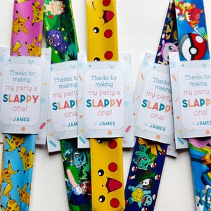 Könnte beinhalten: Mehrere farbenfrohe Schlüsselbänder mit Cartoon-Charakteren, jeweils mit einer Karte versehen. Auf der Karte steht "Danke, dass du meine Party zu einer SLAPPY gemacht hast!" und der Name "JAMES". Die Schlüsselbänder variieren in Farbe und Design.