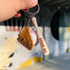 May include: A baseball-themed keychain with a brown leather baseball glove, a white baseball, and a wooden baseball bat with the name "ADAM #9" engraved on it.