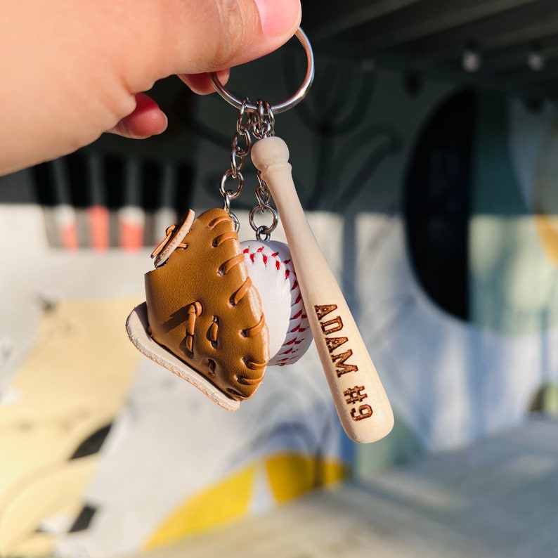 May include: A baseball-themed keychain with a brown leather baseball glove, a white baseball, and a wooden baseball bat with the name "ADAM #9" engraved on it.