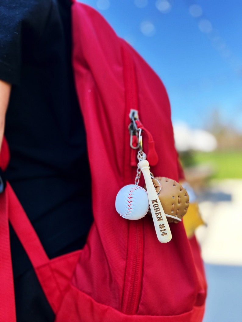 May include: A red backpack with a baseball-themed keychain. The keychain features a miniature baseball, a miniature baseball glove, and a wooden bat with the name "KOHEN #14" engraved on it.