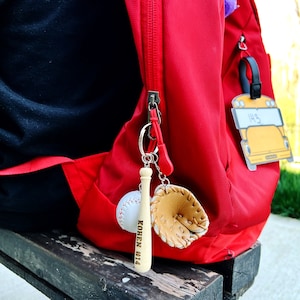 May include: A red backpack with a wooden baseball bat keychain with the text "Kohen #14" engraved on it. The keychain also includes a small leather baseball glove charm.