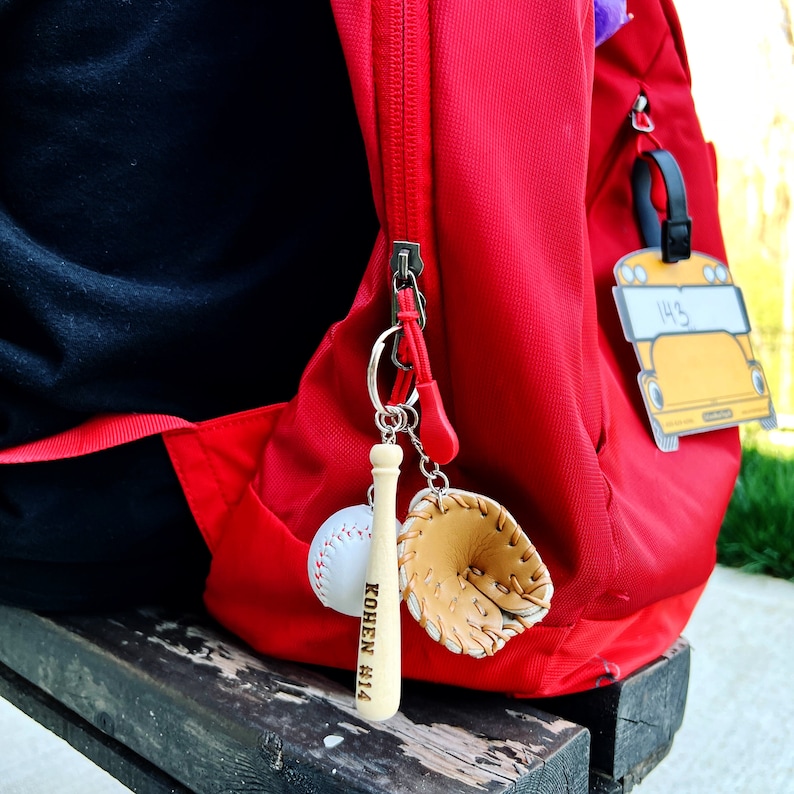 May include: A red backpack with a wooden baseball bat keychain with the text "Kohen #14" engraved on it. The keychain also includes a small leather baseball glove charm.