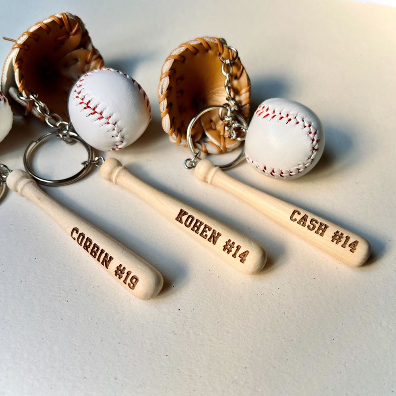 May include: Three wooden baseball bat keychains with personalized names and numbers. The bats are light brown and have the names "Corbin #19", "Kohen #14", and "Cash #14" engraved on them. Each keychain also includes a miniature baseball and a leather baseball glove.