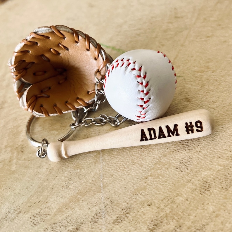 May include: A baseball-themed keychain with a brown leather baseball glove, a white baseball, and a wooden bat with the name "ADAM #9" engraved on it.