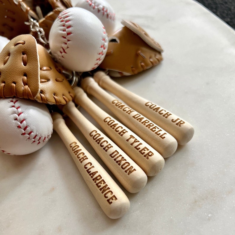 May include: Four wooden baseball bats with the names "Coach Clarence", "Coach Dixon", "Coach Tyler", and "Coach Darrell Jr." engraved on them. The bats are arranged in a row, with the names facing the viewer. The bats are a light brown color and have a smooth finish.