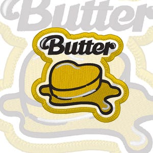 May include: An embroidered patch featuring a yellow heart-shaped stick of butter melting into a puddle with a black outline. The word Butter is stitched in black script font above the heart shape, all set against a white background border.