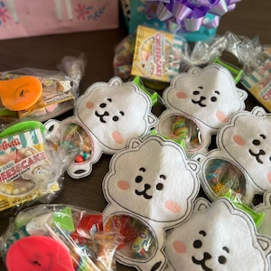 Peut inclure: Une collection de petits sacs en feutre blanc avec des visages de dessins animés brodés. Chaque sac est rempli de bonbons colorés et enveloppé dans du plastique transparent. Les sacs sont disposés sur une surface en bois.