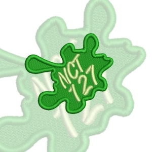 May include: An embroidered patch featuring a bright green irregular blob shape with a dark green border. The text NCT 127 is stitched in a light yellow thread across the center of the blob. The patch is layered over a larger, pale green shape.