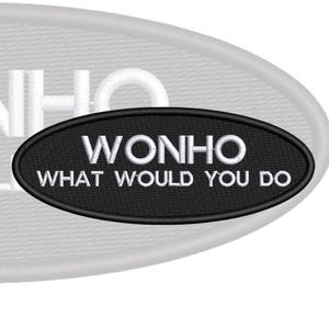 Op de afbeelding: Een ovaalvormige geborduurde patch met een zwarte achtergrond en een zwarte rand. De tekst "WONHO" is in witte hoofdletters op de bovenste regel geborduurd en de tekst "WHAT WOULD YOU DO" is in witte hoofdletters daaronder geborduurd.