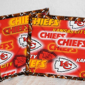 May include: Two square fabric items featuring the Kansas City Chiefs logo and team name in red, white, and yellow. The border is brown with footballs. The items are likely coasters or pot holders.