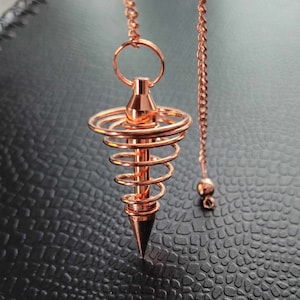 Puede incluir: Un p&eacute;ndulo de metal color cobre con una punta central rodeada por un dise&ntilde;o de espiral de alambre. El p&eacute;ndulo est&aacute; suspendido de una cadena met&aacute;lica a juego que incluye una peque&ntilde;a cuenta esf&eacute;rica en el extremo de la cadena.