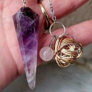 May include: A faceted purple amethyst pendulum point attached to a silver-toned chain. The chain features a small, round, pale pink rose quartz bead and a decorative cluster of intertwined gold, rose gold, and silver-toned wire.
