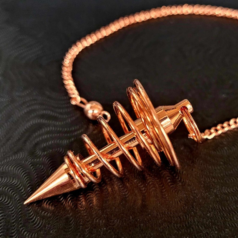 Puede incluir: Un p&eacute;ndulo de metal color oro rosa con una punta afilada y una serie de bobinas de alambre circulares conc&eacute;ntricas envueltas alrededor de una varilla central. El p&eacute;ndulo est&aacute; unido a una cadena de metal a juego con una peque&ntilde;a cuenta esf&eacute;rica.