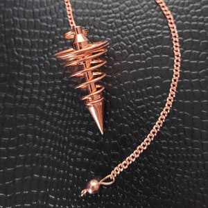 Puede incluir: Un p&eacute;ndulo de metal color cobre con una punta c&oacute;nica y un dise&ntilde;o de alambre en espiral que se estrecha hacia la parte superior. El p&eacute;ndulo est&aacute; unido a una cadena de metal a juego de eslabones finos que termina en una peque&ntilde;a cuenta pulida.