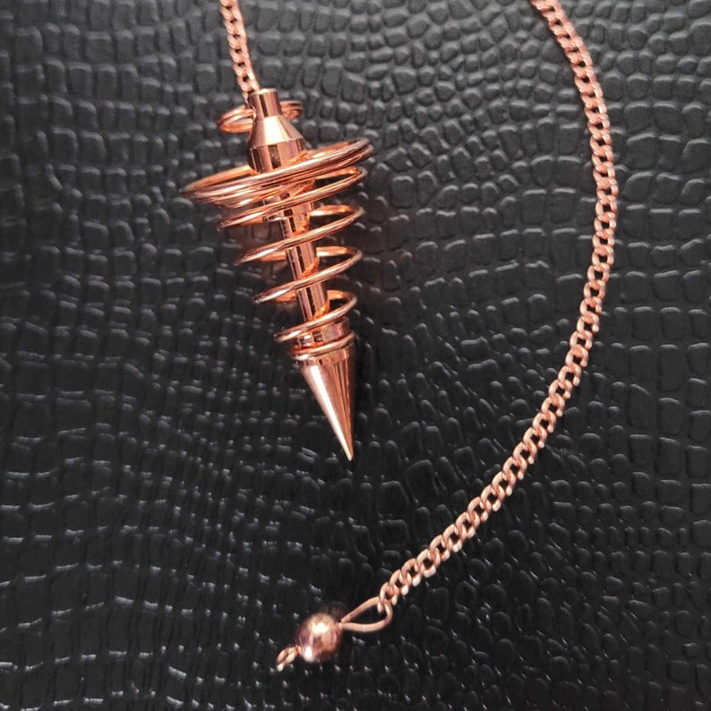 Puede incluir: Un p&eacute;ndulo de metal color cobre con una punta c&oacute;nica y un dise&ntilde;o de alambre en espiral que se estrecha hacia la parte superior. El p&eacute;ndulo est&aacute; unido a una cadena de metal a juego de eslabones finos que termina en una peque&ntilde;a cuenta pulida.