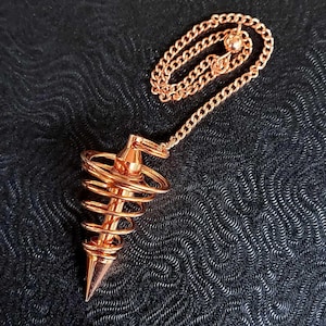 Puede incluir: Un p&eacute;ndulo de metal color cobre con una base c&oacute;nica puntiaguda rodeada por un dise&ntilde;o de espiral de alambre. El p&eacute;ndulo est&aacute; unido a una cadena de eslabones met&aacute;licos a juego que presenta una peque&ntilde;a cuenta esf&eacute;rica en el extremo.