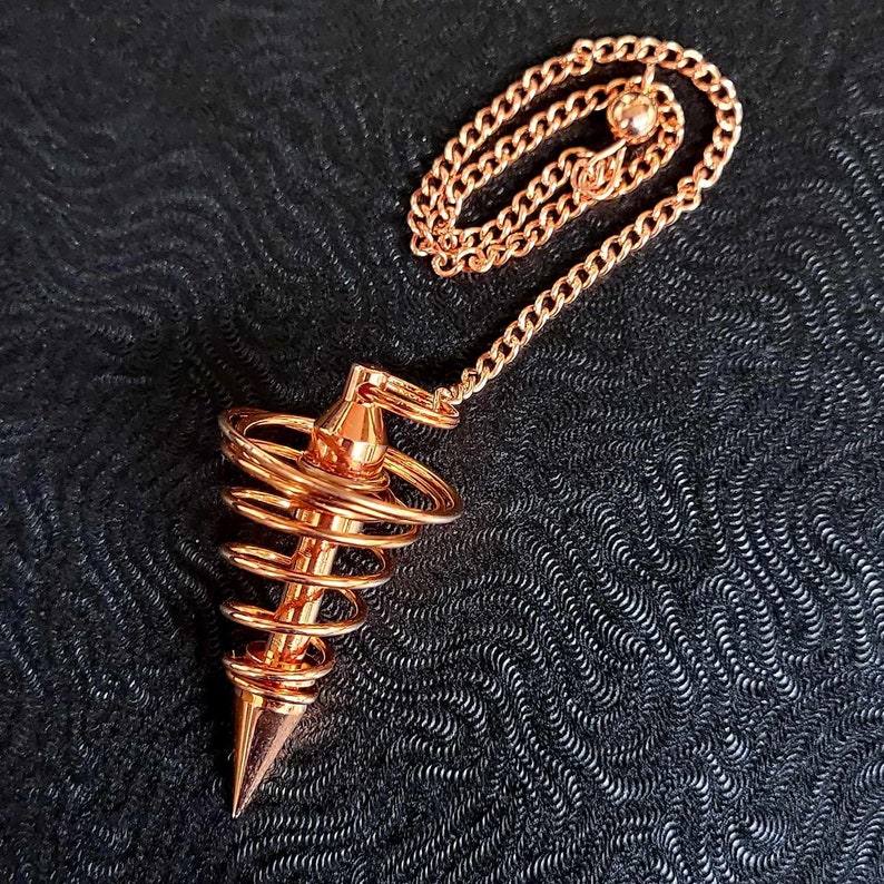 Puede incluir: Un p&eacute;ndulo de metal color cobre con una base c&oacute;nica puntiaguda rodeada por un dise&ntilde;o de espiral de alambre. El p&eacute;ndulo est&aacute; unido a una cadena de eslabones met&aacute;licos a juego que presenta una peque&ntilde;a cuenta esf&eacute;rica en el extremo.