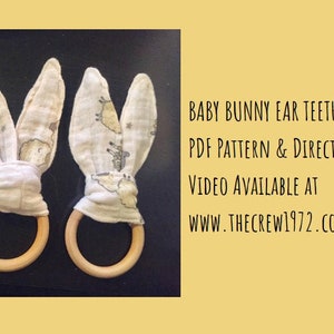 May include: Three wooden teething rings with fabric bunny ears attached. The fabric is white with a light gray pattern of animals and stars. The text on the image reads "Baby Bunny Ear Teether PDF Pattern & Directions Video Available at www.thecrew1972.com"