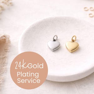 May include: Two heart-shaped pendants, one silver and one gold, rest on a white, textured surface. The pendants have a small loop at the top for attaching to a chain. A circular graphic with the text "24k Gold Plating Service" is in the foreground.