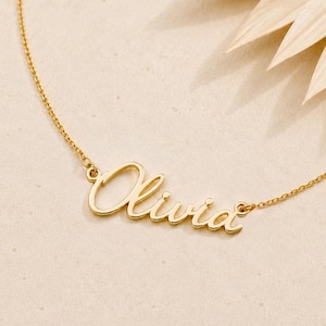 Könnte beinhalten: Eine goldfarbene Halskette mit dem Namen "Olivia" in Schreibschrift. Die Kette liegt auf einer hellen Oberfläche, im Hintergrund ein getrocknetes Palmblatt. Die Kette ist filigran, der Name ist der Blickfang.