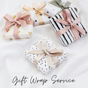 K&ouml;nnte beinhalten: Vier Geschenkboxen, die in verschiedenen Mustern mit rosa und beigefarbenen B&auml;ndern verpackt sind. Die Boxen befinden sich auf einer wei&szlig;en Oberfl&auml;che mit dem Text "Gift Wrap Service" darunter.