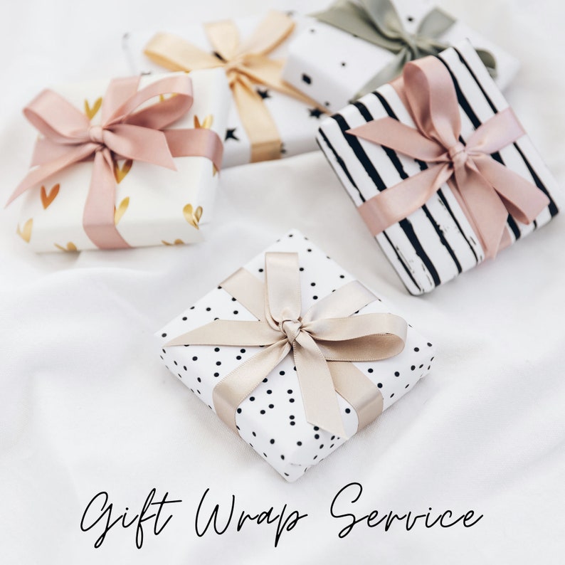 K&ouml;nnte beinhalten: Vier Geschenkboxen, die in verschiedenen Mustern mit rosa und beigefarbenen B&auml;ndern verpackt sind. Die Boxen befinden sich auf einer wei&szlig;en Oberfl&auml;che mit dem Text "Gift Wrap Service" darunter.