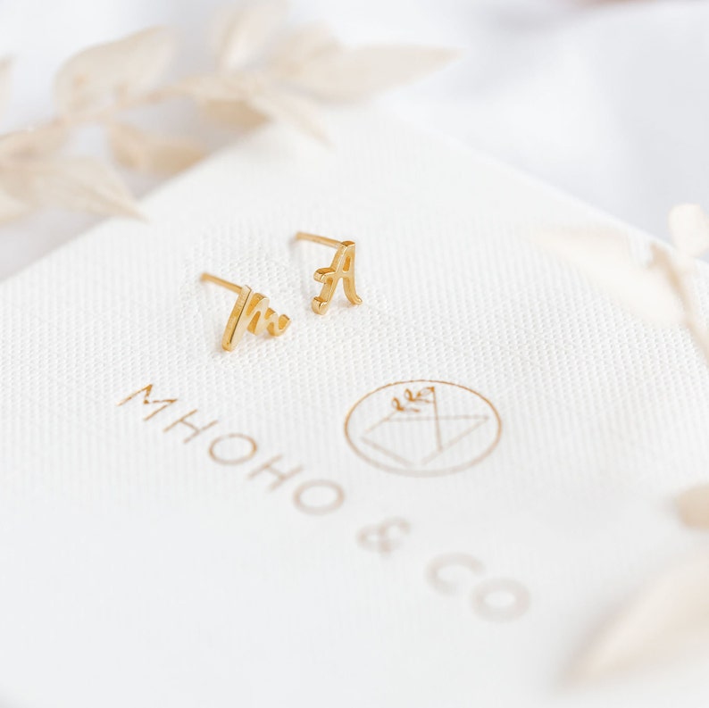 K&ouml;nnte beinhalten: Ein Paar goldene Initialohrringe, einer mit dem Buchstaben "M" und der andere mit dem Buchstaben "A". Die Ohrringe liegen auf einer wei&szlig;en Oberfl&auml;che mit dem aufgedruckten Text "MIHOHO & CO".