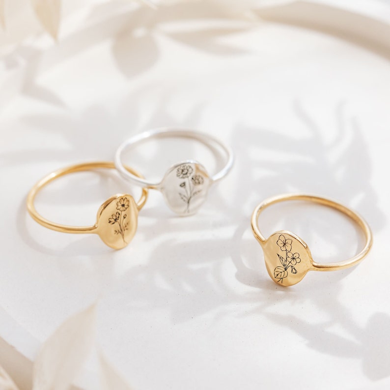 May include: Three delicate gold and silver rings with floral designs. Each ring features a small oval-shaped charm with a simple line drawing of a flower.