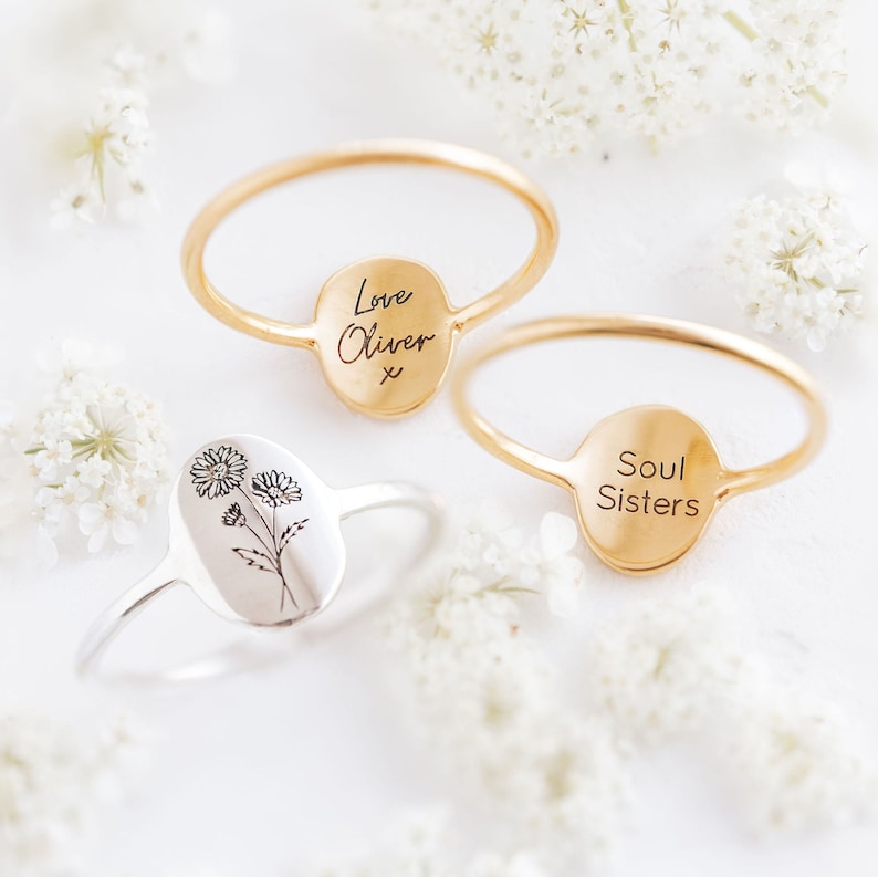May include: Two gold rings with oval-shaped engraved plates. One ring says "Love Oliver x" and the other says "Soul Sisters". A silver ring with an engraved floral design is also shown.