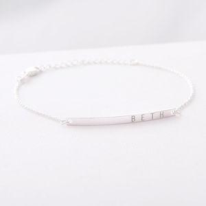 May include: A delicate silver bracelet with a slender, curved bar pendant. The word "BETH" is engraved on the bar. The bracelet features a fine chain and a clasp closure. A simple and elegant piece of jewellery.