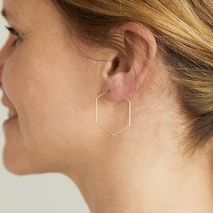 Pode incluir: Um par de brincos de argola hexagonais de ouro. Os brincos são usados por uma pessoa com cabelo comprido e loiro.