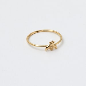 Könnte beinhalten: Ein zarter goldener Ring mit einem kleinen, detaillierten Bienenanhänger. Der Ring hat ein dünnes Band und eine polierte Oberfläche. Die Biene ist das zentrale Element und verleiht dem Ring einen Hauch von von der Natur inspirierten Charme.