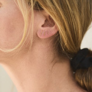 Puede incluir: Primer plano de la oreja de una persona con tres pequeños pendientes de barra de oro. Los pendientes son simples y delicados, y son una opción popular para la joyería minimalista.