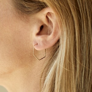 Può includere: Primo piano di un orecchio con un orecchino a cerchio geometrico color oro. L'orecchino ha un design angolare unico, con una forma esagonale. L'orecchino è un accessorio semplice ma elegante.