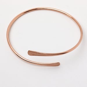May include: A rose gold open cuff bracelet. The bracelet has a simple, minimalist design with a curved, open-ended shape. The ends of the bracelet are flattened. The bracelet is approximately 15 cm in diameter.