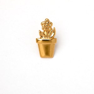 May include: A gold-colored flower pot pin with a detailed flower and leaves. The pin is made of a shiny metal and is set against a white background. The design is simple and elegant.