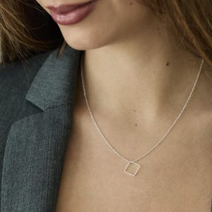 Op de afbeelding: Een zilveren ketting met een vierkante hanger. De hanger is gemaakt van dun draad en heeft een eenvoudig, geometrisch ontwerp.