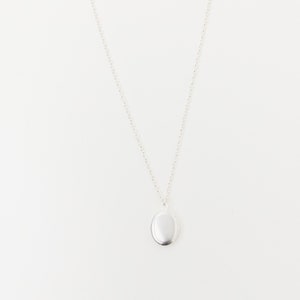 Op de afbeelding: Een zilveren ovale medaillonhanger hangt aan een fijne ketting. Het medaillon is glad en reflecterend, afgebeeld tegen een effen witte achtergrond. De ketting is een eenvoudig, elegant sieraad.