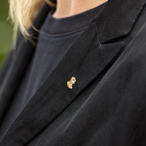 Könnte beinhalten: Eine goldfarbene Anstecknadel in Form eines kleinen Blumen- oder Blätterbüschels ist am Revers eines schwarzen Blazers befestigt. Der Blazer wird über einem schwarzen T-Shirt getragen. Der Hintergrund ist unscharf.