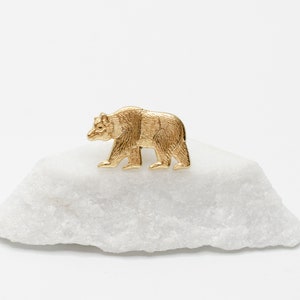 May include: A gold-colored bear figurine, standing on a white, textured rock. The bear is detailed with fur and facial features. The background is plain white, highlighting the bear and rock.